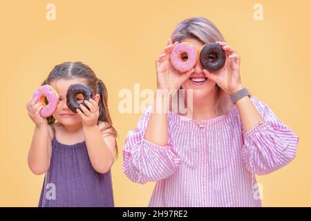 Divertente giovane donna e bambino su sfondo giallo parete. Madre e sua figlia ragazza si divertono con ciambelle colorate. Ciambelle rosa e al cioccolato. Spazzatura Foto Stock