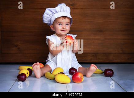 la bambina nel cappello di uno chef è seduta sul pavimento, tenendo una mela e sorridendo. mele, manghi, banane, prugne giacciono sul pavimento accanto a Foto Stock