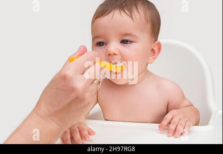 la prima alimentazione del bambino. il bambino apre la bocca per mangiare la purea di arancio naturale da un cucchiaio di arancia dalla mano della madre Foto Stock