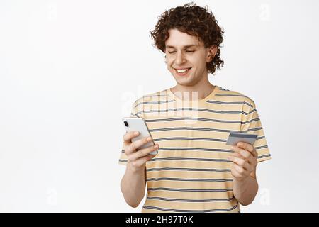 Ritratto di sorridente uomo riccio acquistare online, guardando il cellulare, tenendo la carta di credito, in piedi su sfondo bianco Foto Stock