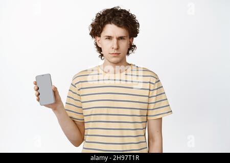 Un ragazzo sospetto che mostra lo schermo del telefono cellulare, squinting e guardando con incredulità la fotocamera, in piedi su sfondo bianco Foto Stock