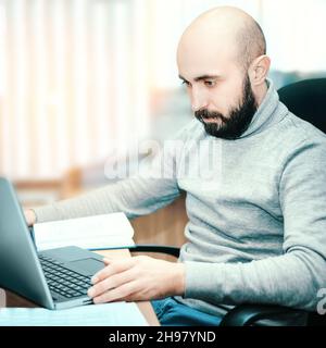 Giovane calvo e barbuto uomo di 25-35 anni lavora in ufficio per computer portatile. Ragazzo in abiti casual guarda il monitor del laptop. Scena reale. Lavoratore reale. Foto Stock