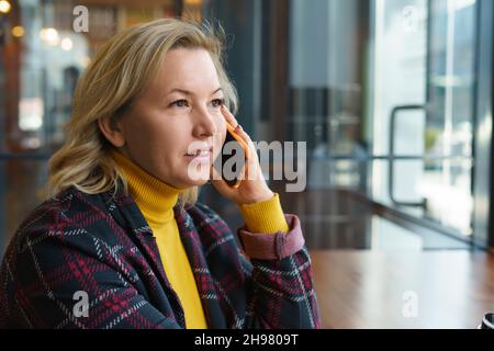 Una bella e positiva bionda caucasica di mezza età sta parlando al telefono cellulare in un bar. Spazio per il testo. Foto Stock