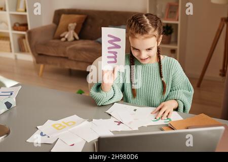 Bambino seduto al tavolo e mostrando la scheda con la lettera durante lo studio online utilizzando il portatile Foto Stock