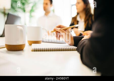 Primo piano di una mano di una donna che tiene una penna e un notebook durante una riunione tra professionisti aziendali nella sala del consiglio d'amministrazione in ufficio Foto Stock