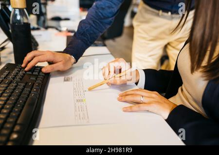 Primo piano delle mani dell'uomo utilizzando il computer del collega e la mano della donna che tiene una penna, su una scrivania disordinata, in ufficio. Lavorare con il toghetere Foto Stock