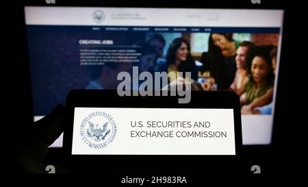 Persona che tiene uno smartphone con il logo della SEC (Securities and Exchange Commission) dell'agenzia statunitense sullo schermo di fronte al sito Web. Mettere a fuoco sul display del telefono. Foto Stock