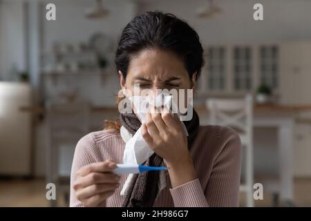 Primo piano donna malata in sciarpa soffia naso, tenendo termometro Foto Stock