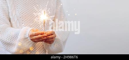 Una ragazza in maglione bianco tiene scintillio bruciante. Festa di Natale di Capodanno concetto. Sfondo chiaro, spazio copia Foto Stock