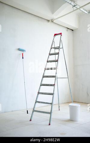 Scala a cavalletto, un secchio di plastica chiuso di vernice e un rullo di vernice, nella stanza. Preparazione per la riparazione. Foto Stock