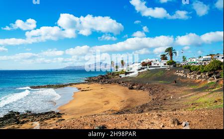 Vista sulla spiaggia di Playa El Barranquillo a Puerto del Carmen, Lanzarote. Spiaggia di sabbia con onde turchesi dell'oceano, case bianche e montagne, Canarie Foto Stock