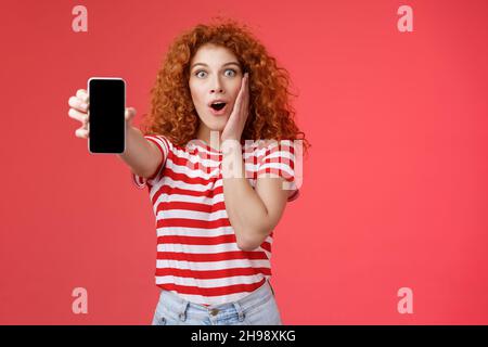 Divertito impressionato redhead buona-osservante ricci-capelli eccitato ragazza condividere impressionante social media pagina mostra smartphone schermo stupito guancia di tocco entusiasta Foto Stock