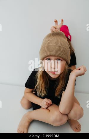 Ragazzo skinny in un cappello di maglia beige cerca la macchina fotografica. Il suo siter gli sta dando orecchie conigliere, nascondendosi dietro di lui Foto Stock