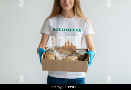 Concetto di aiuto umanitario. Giovane donna in t-shirt e guanti medici volontari, con contenitore per donazioni di cibo, parete luminosa Foto Stock