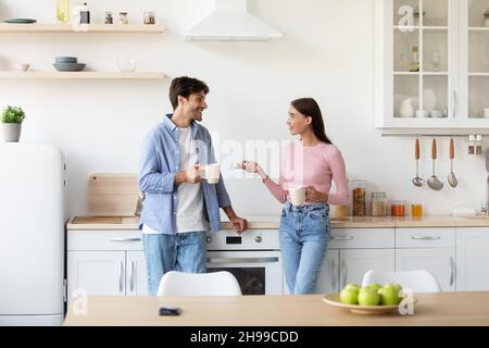 Allegro sorridente giovane moglie e marito con tazze di bevanda calda parlare all'interno della cucina al mattino Foto Stock