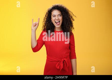 Melomano ribelle che lascia in tenera ragazza. Donna emozionato e spensierata dai capelli ricci in abito rosso che attacca la lingua e guarda bene con il sorriso come Foto Stock