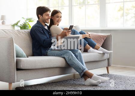 Felice uomo e donna allegro e millenario europeo seduto sul divano con un computer portatile e guardando lo smartphone Foto Stock