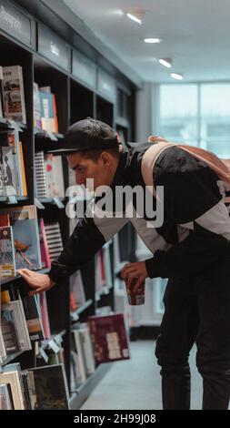 Un giovane uomo adulto che tiene una tazza di caffè e cerca libri in un bibliotecario Foto Stock