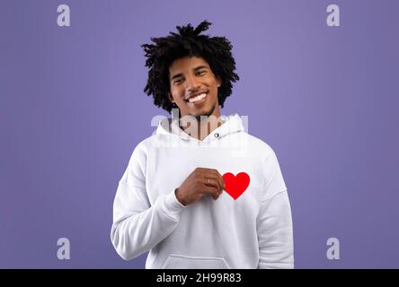 Esprimere affetto. Ragazzo teen nero positivo che tiene piccolo cuore di carta rosso su sfondo viola studio Foto Stock