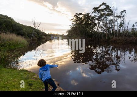 Un giovane ragazzo in piedi accanto ad un fiume al crepuscolo, il cielo si riflette nell'acqua piatta e tranquilla Foto Stock