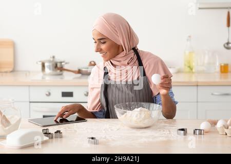 Donna musulmana che verifica la ricetta in linea sulla tavoletta digitale mentre cuoce in cucina Foto Stock