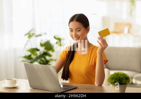 Semplice concetto di pagamento contactless online. Eccitata donna asiatica che effettua acquisti su laptop e con carta di credito Foto Stock