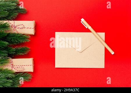 Mockup di busta di carta artigianale e penna artigianale su sfondo rosso con decorazioni di rami di albero di Natale e scatole regalo Foto Stock