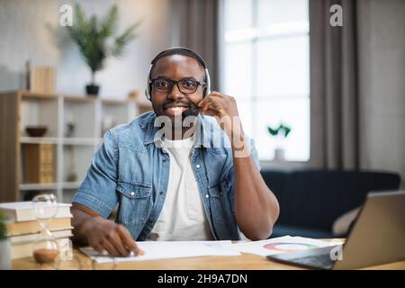 Giovane ragazzo africano sorridendo e guardando la macchina fotografica mentre si siede a casa con un moderno computer portatile. Buon freelance che indossa le cuffie durante le chiamate in conferenza. Foto Stock