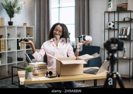 Influencer femminile afroamericana che registra video sulla telecamera mentre si apre la scatola con occhiali VR e joystick. Concetto di blogging e tecnologia moderna. Foto Stock