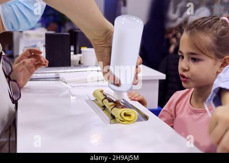 Una bambina aspetta un pasticciere per versare la panna bianca montata sulle frittelle. Fast food. Primo piano, messa a fuoco selettiva Foto Stock