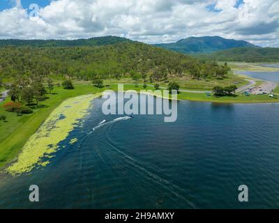 Un motoscafo che tira due persone su tubi gonfiabili toglie dalle rive una grande diga fiancheggiata da piante acquatiche Foto Stock