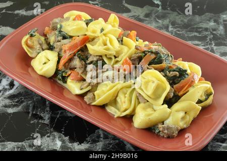 Tortellini al parmigiano con spinaci, bistecche e pomodori cena su piatto rosso Foto Stock