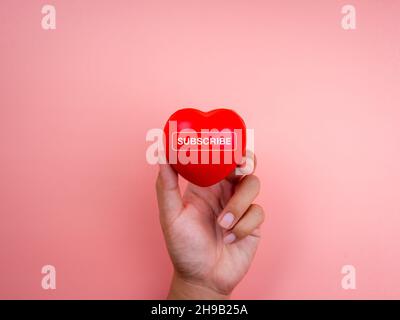 Concetto di sottoscrizione. Parola 'SUBSCRIBE' su palla rossa a cuore in mano su sfondo rosa pastello con spazio copia, stile minimal. Dopo i social media, acquisto Foto Stock