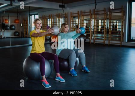 Donna sovrappeso con manubri seduti su palla fintess e allenarsi con personal trainer in palestra Foto Stock