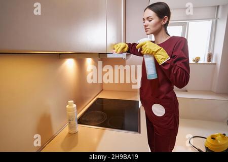 Calmo janitor femminile messo a fuoco in guanti di gomma pulizia cappa di scarico appeso sopra piano di cottura Foto Stock