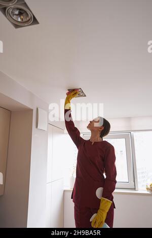 Janitor focalizzato in guanti di gomma strofinando i dispositivi di illuminazione da soffitto incassati con panno in microfibra Foto Stock