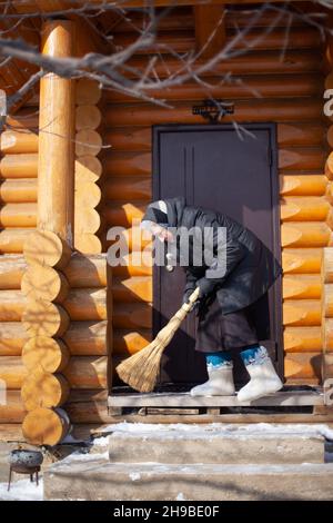 La donna anziana spazza la neve dal portico. Donna caucasica in abiti neri, stivali in feltro e sciarpa rimuove la neve dal portico con scopa, che cadde dopo Foto Stock