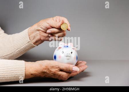 La mano rugosa di una donna anziana mette una moneta in euro in una banca piggy. Sfondo grigio. Il concetto di risparmio e pensionamento. Foto Stock