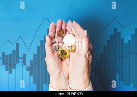 Le mani di femakes rugose tengono una manciata di bitcoins e monete d'oro. Business chart blu sullo sfondo. Il concetto di banca e di investimento. Foto Stock