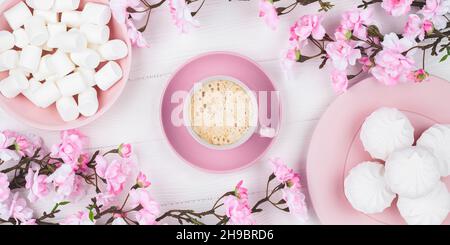 Romantica primavera ancora vita. Tazza di caffè con marshmallow al mattino per colazione. Fiore di ciliegio su sfondo bianco in legno. Disposizione piatta. Rosa pastello Foto Stock