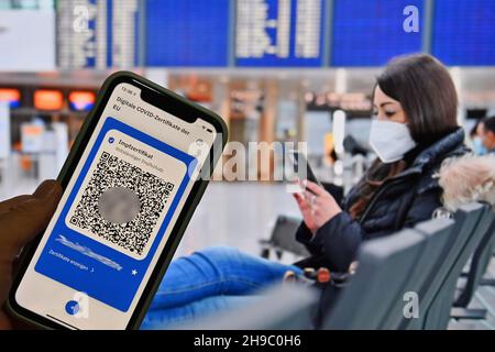 Foto topica/simbolo: Certificati digitali del Covid dell'UE su smartphone, protezione completa della vaccinazione, certificato di vaccinazione, tessera di vaccinazione digitale, tessera di vaccinazione. Giovane donna con maschera facciale, maschera con valigia, bagaglio all'aeroporto di Monaco il 6 dicembre 2020. Seduto al terminal, sala partenze, in attesa di partenza e guardando il suo smartphone. MODELLO RILASCIATO! Foto Stock