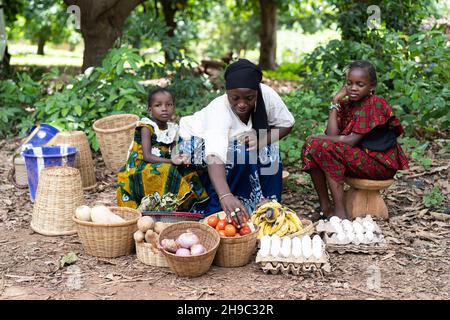 Black West African Street market venditore che mostra i suoi prodotti casalinga seduto sul bordo di una strada del villaggio con due delle sue figlie Foto Stock