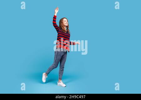 Ritratto a tutta lunghezza di donna sorridente felice in maglione, stand con braccio sollevato, guardando a distanza con sorriso affascinante, esprime emozioni positive. Studio interno girato isolato su sfondo blu. Foto Stock