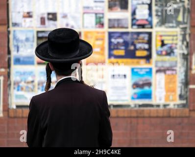 Un ebreo asidico con un lungo peyot ricurly legge annunci e pubblicità su un muro della comunità a Williamsburg, Brooklyn, New York City. Foto Stock