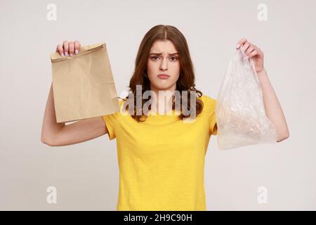 Giovane ragazza adolescente sconvolto in giallo casual t-shirt tenendo due borse in mano, carta e plastica, guardando la tristezza della fotocamera, rifiutando di utilizzare la plastica. Studio interno girato isolato su sfondo grigio. Foto Stock