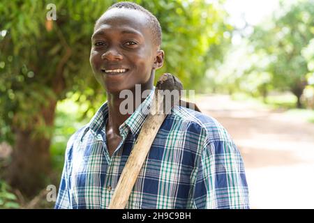 Bel contadino nero afro-americano in vestito casual con una zappa sulle spalle sorridendo con fiducia alla macchina fotografica Foto Stock