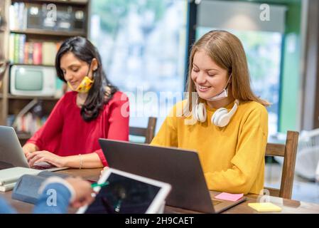 Due giovani donne che studiano in biblioteca insieme, preparandosi per gli esami. Studenti universitari multietnici. Due studenti universitari che studiano con facemask Foto Stock