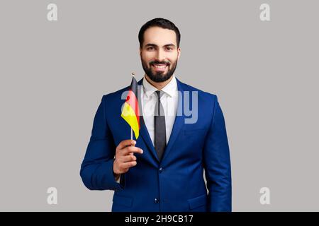 Felice uomo d'affari che detiene la bandiera della Germania, celebrando il giorno della Germania - 3 ottobre, esprimendo emozioni positive, indossando abiti stile ufficiale. Studio interno girato isolato su sfondo grigio. Foto Stock
