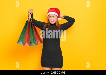 Bella giovane donna asiatica in Babbo Natale cappello che tiene borse shopping e testa graffiante con mano isolato su sfondo giallo Foto Stock