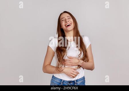 Donna estremamente felice tenendo lo stomaco e ridendo fuori forte, ridacchiando ridacchiando all'aneddoto divertente, emozione sincera, indossando T-shirt bianca. Studio interno girato isolato su sfondo grigio. Foto Stock